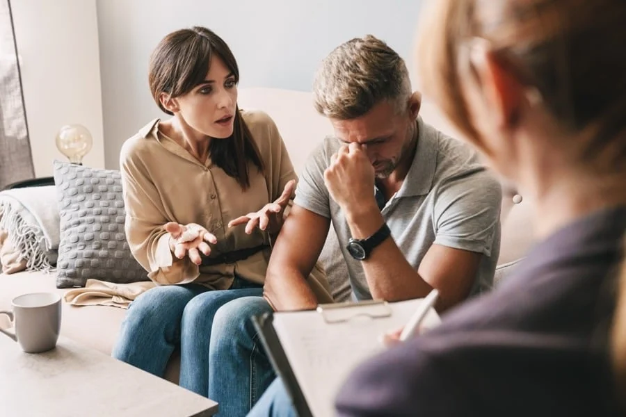supporting-alcoholic-partner Woman argues with frustrated man sitting next to her on couch while therapist holding clipboard looks on