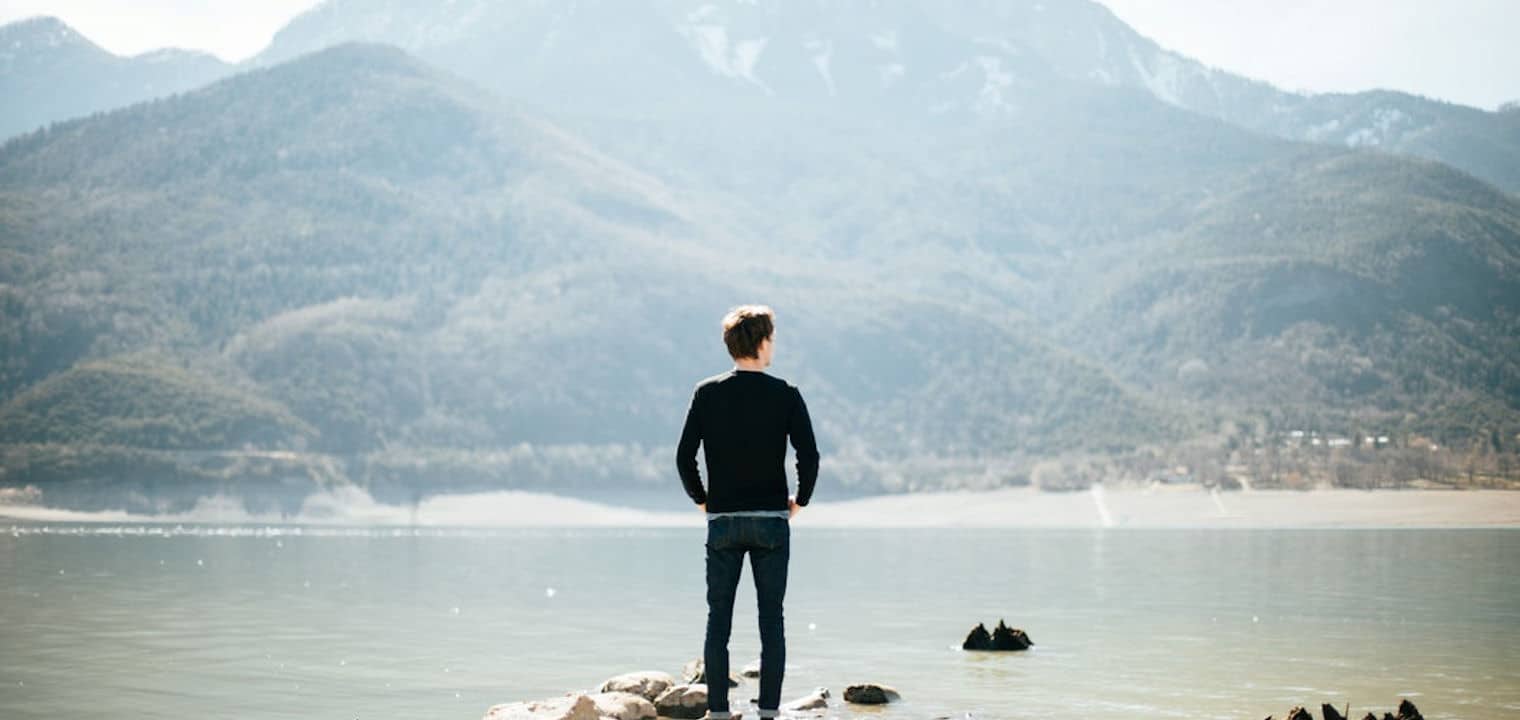 Man looking out from rocky shore onto a lake in front of a mountain
