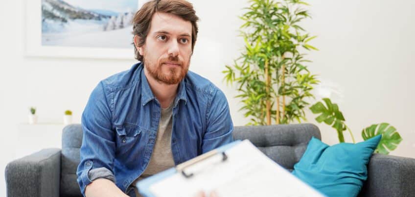 Man sitting on couch eagerly waiting to hear results on clipboard