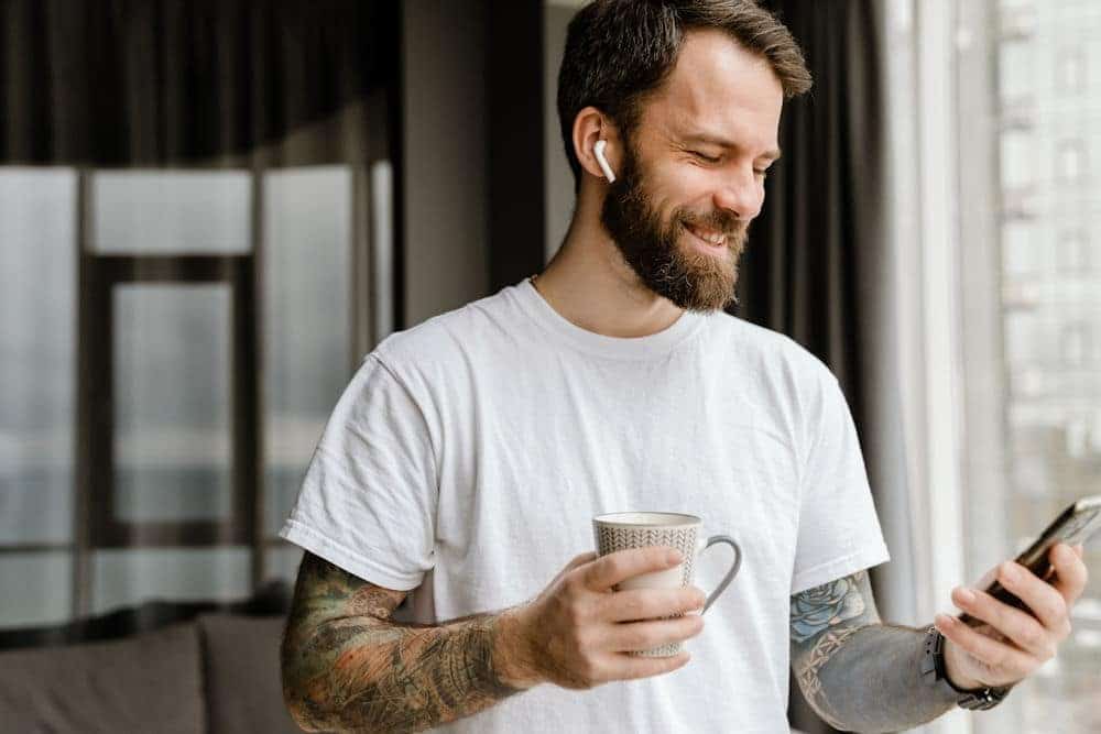 Cheerful man with tattoo sleeves smiles at smartphone while holding coffee and wearing earbud