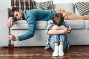 Sad child sitting on floor while alcoholic parent sleeps on couching holding a handle of whiskey