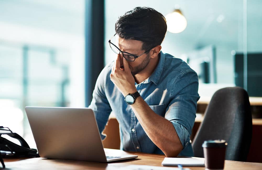 Man with anxiety pinching bridge of his nose while looking down at laptop