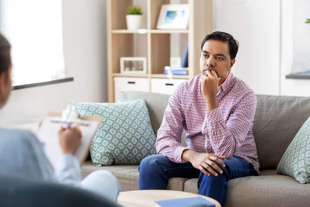 Man sitting on couch learns about signs of alcoholism from therapist with clipboard