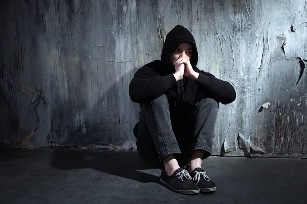 Young man in hoodie thinks while sitting against wall and clasping hands