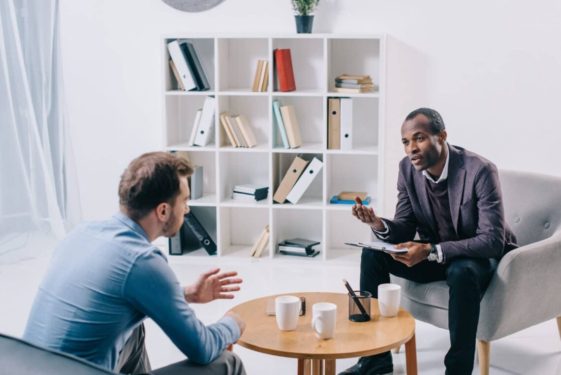 Man sitting down with therapist in mostly white office.