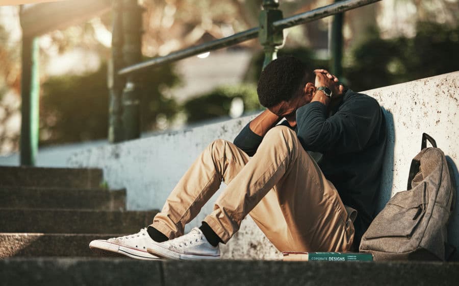 Man with backpack braces neck with hands while sitting against wall on concrete steps.