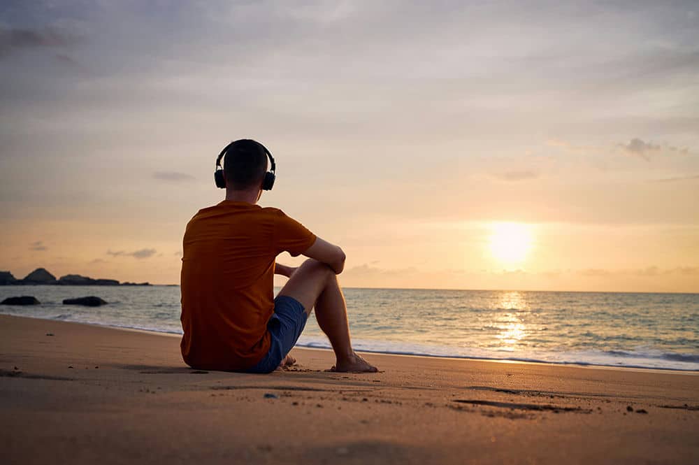 Man sitting on the beach wearing cans-style headphones while contemplating personal trauma.