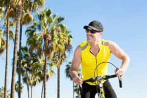 Man in yellow racing shirt and black baseball cap pauses riding bike down road with palm trees.